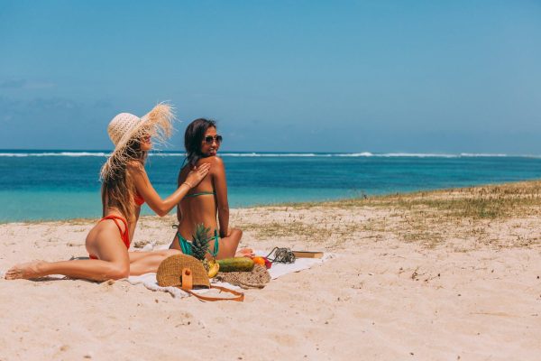 A woman in a red bikini and a hat is applying sunscreen to a woman in a green bikini and sunglasses. cruise packing list for women