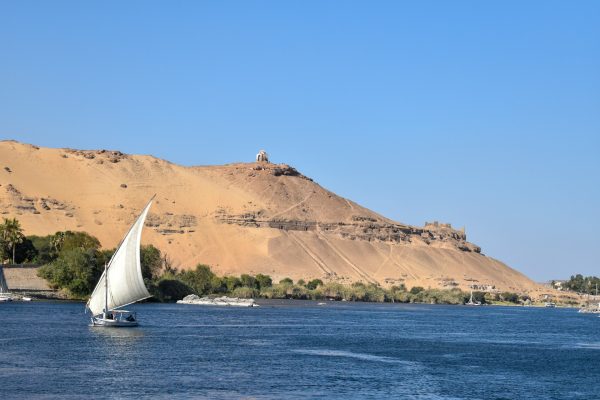 Traditional felucca sailing on the Nile River with the Tombs of the Nobles on the hillside in Aswan, Egypt